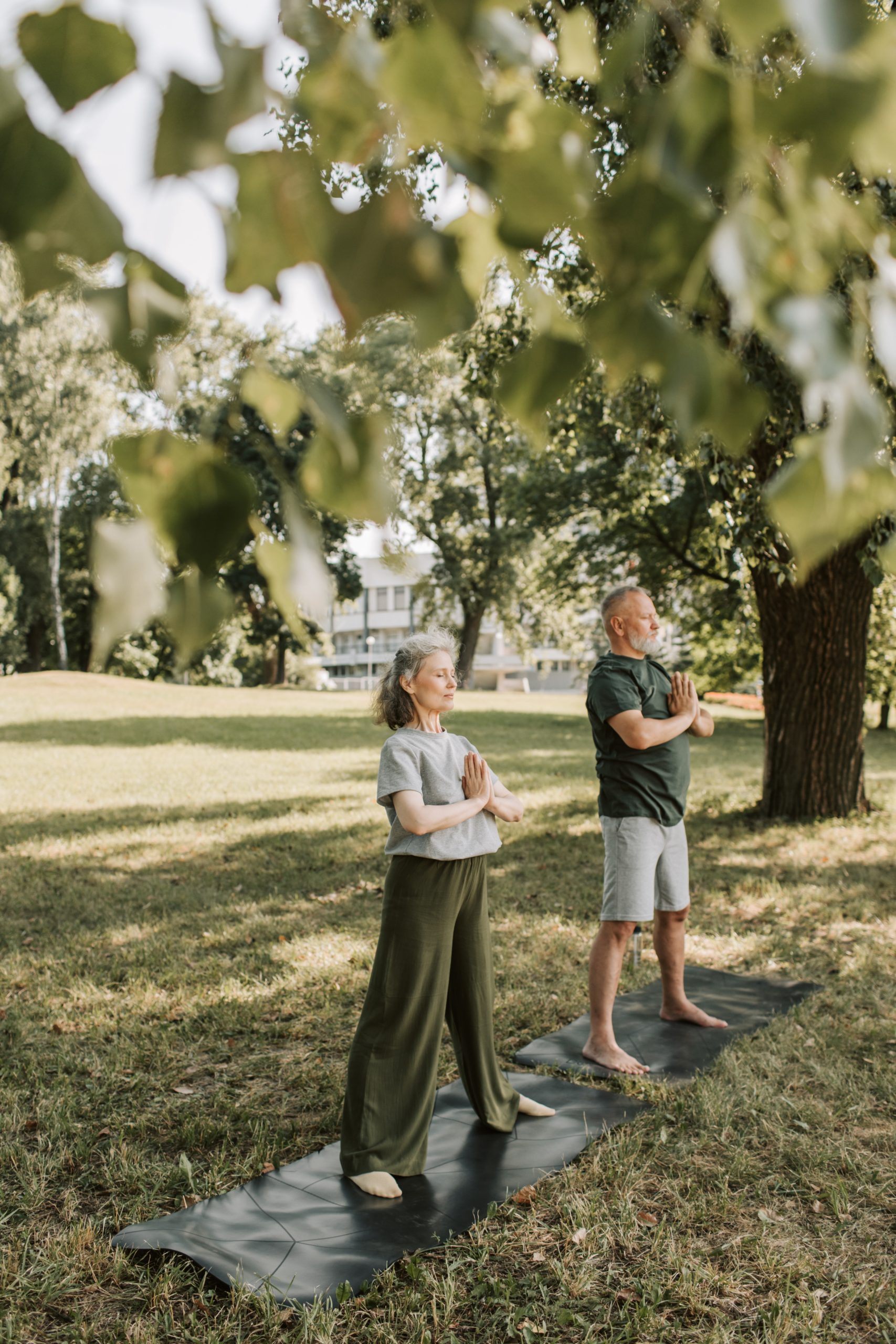 A woman with grey hair and a man with grey hair on a yoga mat outdoors under a tree.
