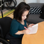 A South Asian person in her wheelchair takes notes by hand during a meeting.