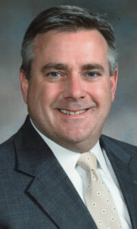 Photo of male with dark gray hair, smiling, and wearing a gray suit and tan tie.