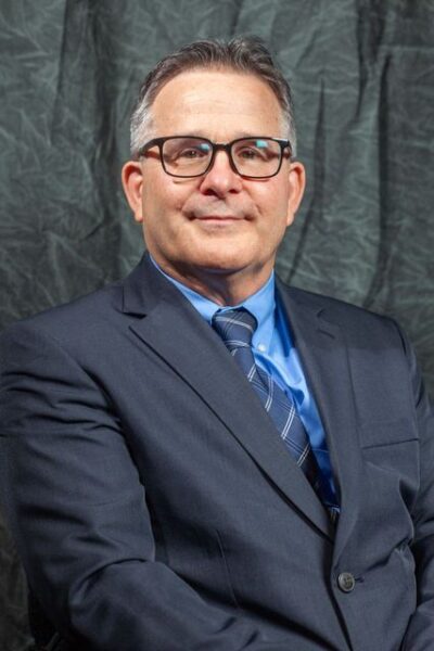 Gray Background. Professional headshot of male with glasses, wearing a dark blue jacket, light blue shirt and multi-blue tie.