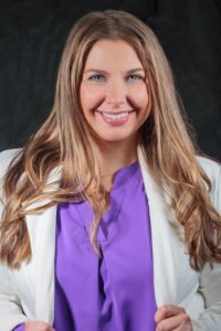 Gray Background. Headshot of female with long brown hair, smiling, wearing a purple shirt and white blazer.