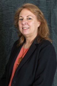 Gray Background. Professional headshot of female smiling, with shoulder-length auburn hair, wearing a pink shirt and black jacket.