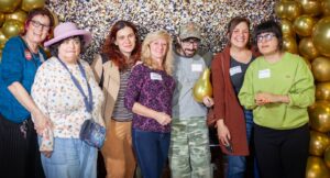 Image Description: Group of individuals from the Home Care Mixer Event, including Personal Aides and Consumers, standing in front of a black and gold banner, smiling and looking happy to be with their friends