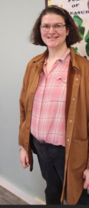 Photo of Jen, wearing a pink shirt and brown jacket, her short brown hair pushed back with a headband. She is smiling in front of a bulletin board.