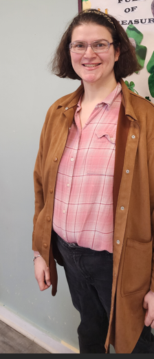 Photo of Jen, wearing a pink shirt and brown jacket, her short brown hair pushed back with a headband. She is smiling in front of a bulletin board. Community Independence Program (CIP)