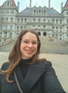 Image of a smiling female with long brown hair, wearing glasses, a cross body purse and a black pea coat. She is taking a selfie in front of the steps of the State Capitol building, a building full of windows, pointed gables and red roofing. 