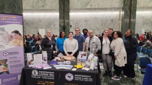 Photo of a passionate group of Independent Living staff, behind the Independent Living table, aside the signage, smiling