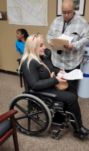 Picture of a female, blonde with glasses on her head, holding a stack of papers and using a wheelchair. She is advocating for the rights of people with disabilities with a legislator.