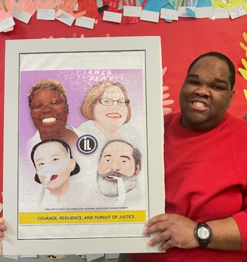 Image of a male consumer wearing a red sweater, holding a framed puzzle that he put together himself. developmental disabilities awareness