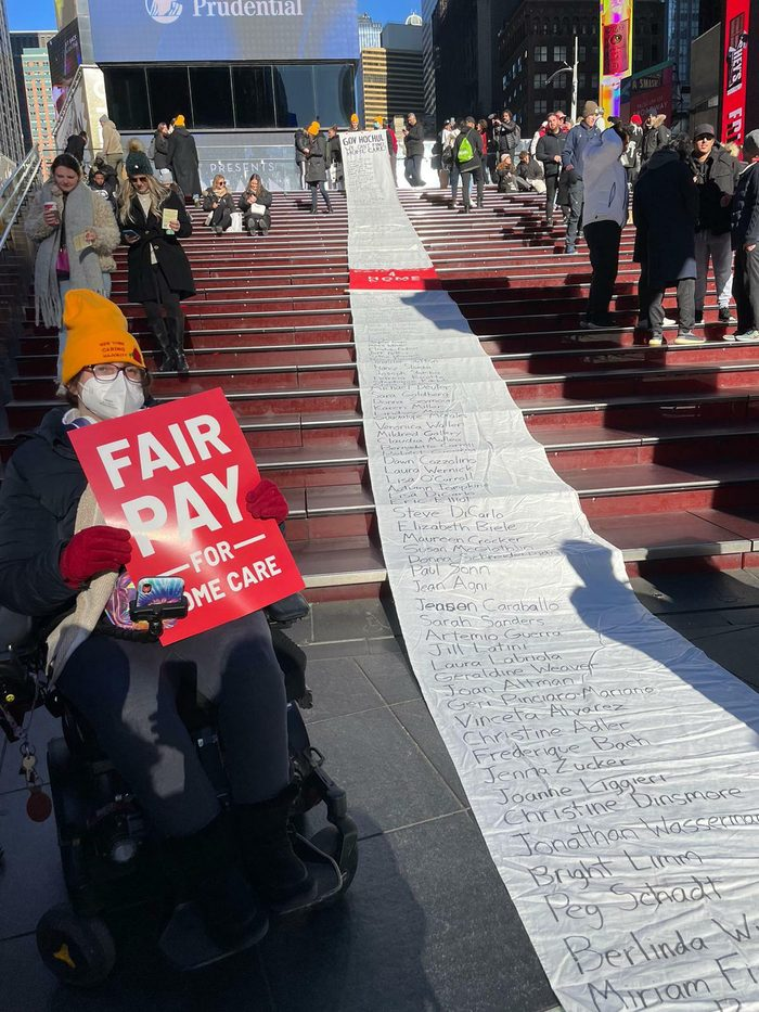 Photo of people lining both sides of a long set of stairs, using wheelchairs, some standing on the stairs, holding signs for Fair Pay For Home Care. Between the people, top the top of the stairs, is a scroll o fnames of people impacted by home care cuts.
