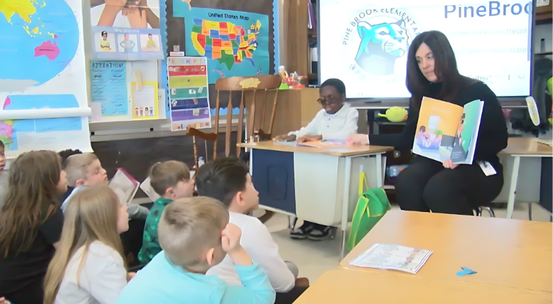 A classroom of children sit on the floor looking up at a teacher reading a picture book created by young child. The child author sits at a small wooden desk at the front of the class.