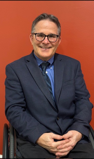 Doug 2025-1 Gray Background. Professional headshot of male with glasses, wearing a dark blue jacket, light blue shirt and multi-blue tie.
